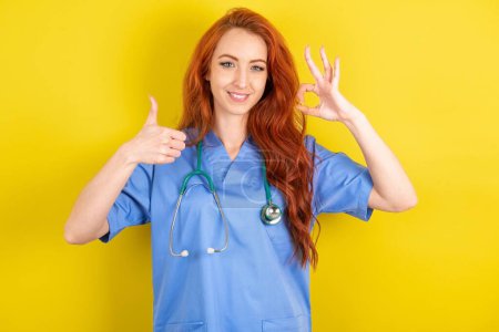 Téléchargez les photos : Jeune femme médecin aux cheveux roux sur fond jaune studio souriant et regardant heureux, insouciant et positif, geste de victoire ou de paix avec une main - en image libre de droit