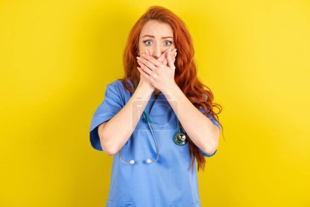 Téléchargez les photos : Stupéfait jeune femme médecin aux cheveux roux sur fond jaune studio couvre les deux mains sur la bouche, appréhension de quelque chose d'étonnant - en image libre de droit