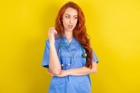 Téléchargez les photos : Étonné jeune femme médecin aux cheveux roux sur fond jaune studio regarde de côté étonnamment avec bouche ouverte. - en image libre de droit
