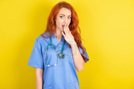 Téléchargez les photos : Jeune femme médecin aux cheveux roux sur fond jaune studio couvre la bouche et regarde avec émerveillement la caméra, ne peut pas croire les rumeurs inattendues. - en image libre de droit