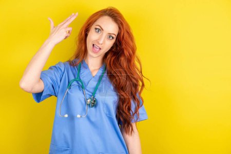 Photo for Young red-haired doctor woman over yellow studio background foolishness around shoots in temple with fingers makes suicide gesture. Funny model makes finger gun pistol - Royalty Free Image