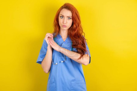 Téléchargez les photos : Surpris émotionnel jeune femme médecin aux cheveux roux sur fond jaune studio frotte les paumes et regarde la caméra avec incrédulité - en image libre de droit