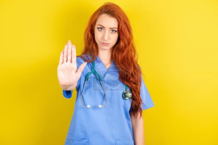 Photo for Young red-haired doctor woman over yellow studio background shows stop sign prohibition symbol keeps palm forward to camera with strict expression - Royalty Free Image