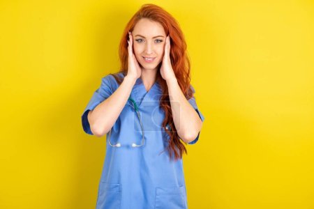 Photo for Happy young red-haired doctor woman over yellow studio background touches both cheeks gently, has tender smile, shows white teeth, gazes positively straightly at camera, - Royalty Free Image