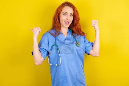 Téléchargez les photos : Hourra cool jeune rousse médecin femme sur jaune studio fond point retour vide espace poing main - en image libre de droit