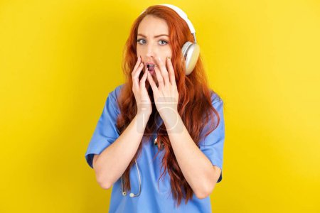 Téléchargez les photos : Choqué jeune femme médecin aux cheveux roux sur fond jaune studio fixe craintif à la caméra garde la bouche largement ouverte porte des écouteurs stéréo sans fil sur les oreilles - en image libre de droit