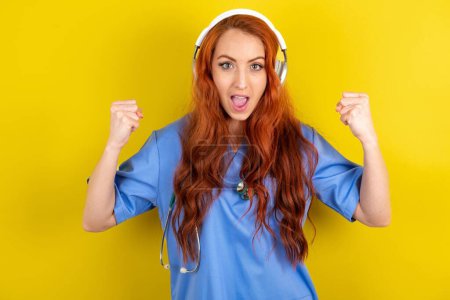 Photo for Emotional young red-haired doctor woman over yellow studio background exclaims loudly feels like winner raises clenched fists keeps mouth opened wears stereo headphones on ears makes yes gesture, listens favourite music - Royalty Free Image