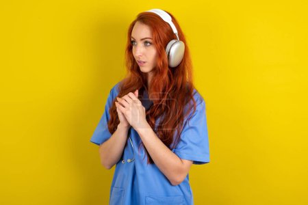 Photo for Young red-haired doctor woman over yellow studio background  wears stereo headphones listening to music concentrated and looking aside with interest. - Royalty Free Image