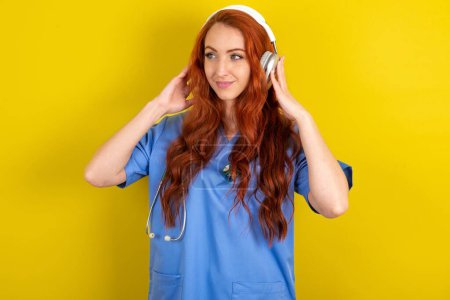 Téléchargez les photos : Jeune médecin rousse femme sur fond jaune studio porte casque stéréo écoute de la musique concentrée de côté. Les gens hobby concept de style de vie - en image libre de droit