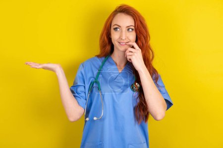 Foto de Positivo joven médico de pelo rojo mujer sobre fondo amarillo estudio anuncio promo toque dientes de dedo - Imagen libre de derechos