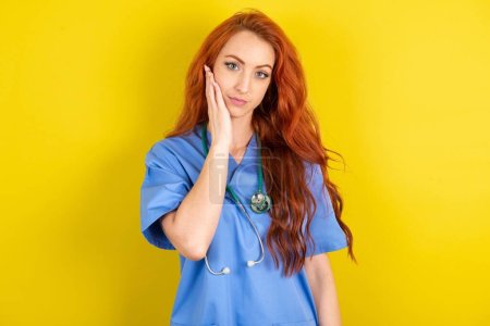 Téléchargez les photos : Photo de jeune femme médecin aux cheveux roux sur fond jaune studio profiter d'une peau fraîche parfaite toucher visage - en image libre de droit