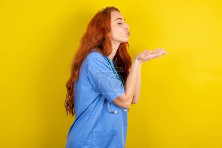 Téléchargez les photos : Profil vue de côté, portrait d'une jolie jeune femme médecin aux cheveux roux sur fond de studio jaune envoyant un baiser d'air - en image libre de droit