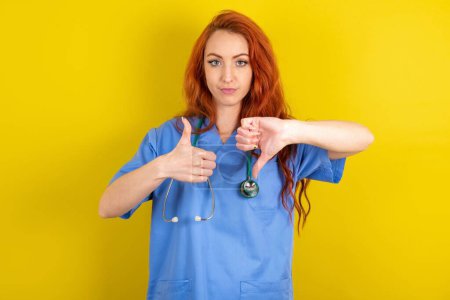 Téléchargez les photos : Jeune rousse médecin femme sur fond jaune studio montrant pouce vers le haut signe vers le bas - en image libre de droit