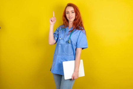 Téléchargez les photos : Jeune femme médecin aux cheveux roux sur fond jaune studio tenant ordinateur portable montrant copie espace vide annonce - en image libre de droit