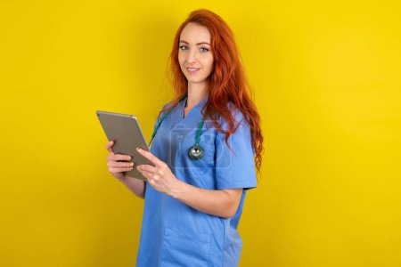 Téléchargez les photos : Photo de jeune femme médecin aux cheveux roux optimiste sur fond jaune studio tenant la tablette - en image libre de droit