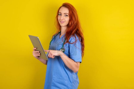Foto de Foto de feliz alegre inteligente joven médico de pelo rojo mujer sobre fondo estudio amarillo celebrar tableta de navegación por Internet - Imagen libre de derechos
