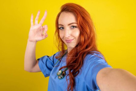 Téléchargez les photos : Portrait de jolie et joyeuse femme aux cheveux roux médecin sur fond jaune studio faire selfie spectacle okey symbole - en image libre de droit