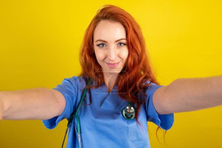 Foto de Foto de una joven doctora pelirroja sobre fondo de estudio amarillo hacer selfie - Imagen libre de derechos