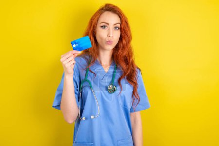 Photo for Photo of young red-haired doctor woman over yellow studio background amazed shocked hold credit card payment - Royalty Free Image