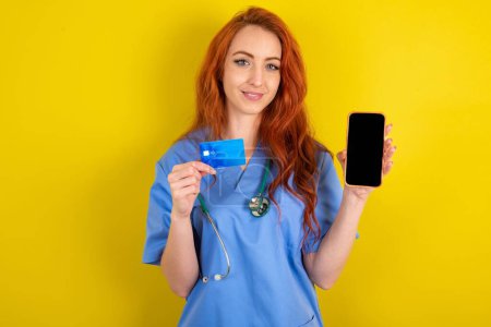 Photo for Photo of adorable young red-haired doctor woman over yellow studio background  holding credit card and Smartphone. Reserved for online purchases - Royalty Free Image