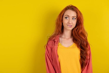 Téléchargez les photos : Incroyable belle femme aux cheveux rouges debout sur fond jaune studio lèvre mordante et l'air délicat à vide espace. - en image libre de droit