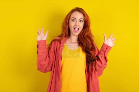Téléchargez les photos : Optimiste belle femme aux cheveux rouges debout sur fond jaune studio soulève les paumes de joie, heureux de recevoir un cadeau impressionnant de quelqu'un, crie bruyamment, - en image libre de droit