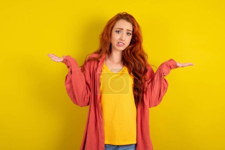 Photo for Clueless beautiful red haired woman standing over yellow studio background shrugs shoulders with hesitation, faces doubtful situation, spreads palms, Hard decision - Royalty Free Image