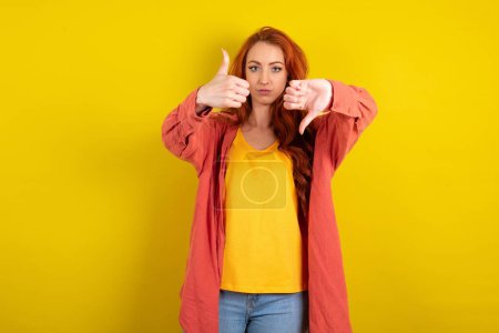 Photo for Beautiful red haired woman standing over yellow studio background showing thumbs up and thumbs down, difficult choose concept - Royalty Free Image