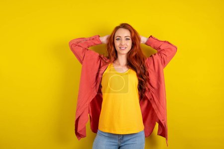 Photo for Satisfied red haired woman standing over yellow studio background hold hands behind head relaxing - Royalty Free Image