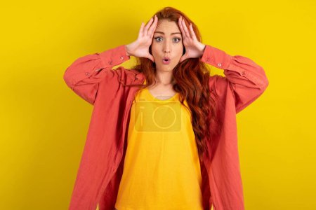 Photo for Beautiful red haired woman standing over yellow studio background with scared expression, keeps hands on head, jaw dropped, has terrific expression. Omg concept - Royalty Free Image