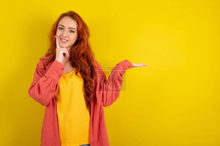 Téléchargez les photos : Funny femme aux cheveux rouges debout sur fond jaune studio tenir palme ouverte nouveau produit grande proposition - en image libre de droit