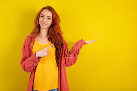 Téléchargez les photos : Positif femme aux cheveux rouges debout sur fond jaune promoteur index de point copyspace main tenir démontrer offre annonces promo - en image libre de droit