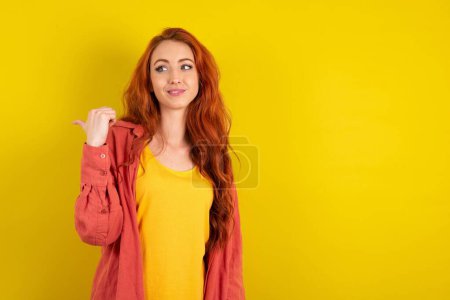 Photo for Charming red haired woman standing over yellow studio background looking at copy space having advertisements - Royalty Free Image