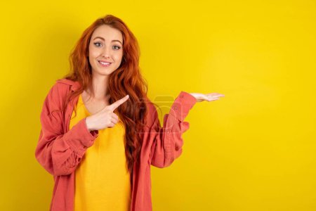 Foto de Hermosa mujer de pelo rojo de pie sobre fondo amarillo estudio señalando y sosteniendo la mano mostrando anuncios - Imagen libre de derechos