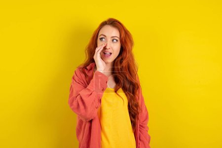 Téléchargez les photos : Belle femme aux cheveux rouges debout sur fond jaune studio entendre incroyable nouvelles privées impressionné partager cri - en image libre de droit