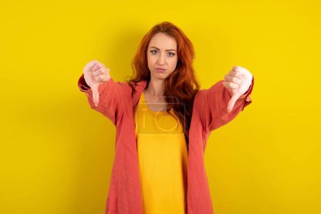 Photo for Beautiful red haired woman standing over yellow studio background being upset showing thumb down with two hands. Dislike concept. - Royalty Free Image