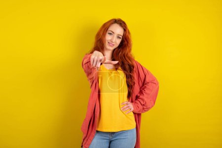 Foto de Hermosa mujer de pelo rojo de pie sobre el fondo del estudio amarillo apuntando a la cámara con una sonrisa satisfecha, confiada, amable, eligiéndote - Imagen libre de derechos