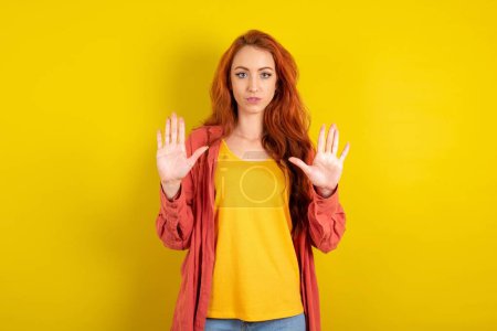 Photo for Serious red haired woman standing over yellow studio background pulls palms towards camera, makes stop gesture, asks to control your emotions and not be nervous - Royalty Free Image