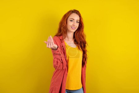 Photo for Beautiful red haired woman standing over yellow studio background inviting to come with hand. Happy that you came - Royalty Free Image