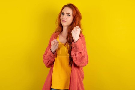 Téléchargez les photos : Belle femme aux cheveux rouges debout sur fond jaune studio Prêt à se battre avec geste de défense poing, visage en colère et contrarié, peur du problème. - en image libre de droit