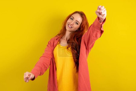 Foto de Hermosa mujer de pelo rojo de pie sobre fondo amarillo estudio imaginar timón timón timón pasar examen de conducción buen humor velocidad rápida - Imagen libre de derechos