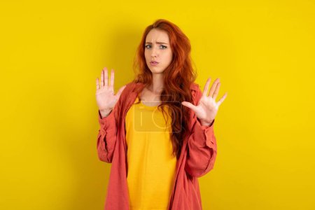 Téléchargez les photos : Belle femme aux cheveux roux debout sur fond jaune studio Déménagement mains paumes montrant refus et déni avec expression effrayante et dégoûtante. Arrêt et interdit. - en image libre de droit
