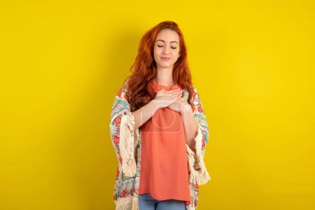 Photo for Red haired woman standing over yellow studio background smiling with hands on chest with closed eyes and grateful gesture on face. Health concept. - Royalty Free Image