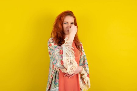 Téléchargez les photos : Femme aux cheveux roux debout sur fond jaune studio sentant quelque chose de puant et dégoûtant, odeur intolérable, retenant souffle avec les doigts sur le nez. Mauvaise odeur - en image libre de droit