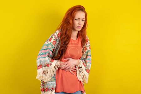 Téléchargez les photos : Femme aux cheveux roux debout sur fond jaune studio avec la main sur le ventre parce que les nausées, la maladie douloureuse se sentent mal. Concept Ache. - en image libre de droit