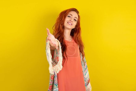 Téléchargez les photos : Femme aux cheveux roux debout sur fond jaune studio souriant amical offrant poignée de main comme accueil et accueil. Une entreprise prospère. - en image libre de droit