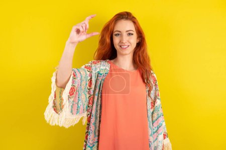 Foto de Mujer de pelo rojo de pie sobre fondo amarillo estudio sonriente y seguro gesto con la mano haciendo signo de tamaño pequeño con los dedos mirando y la cámara. Concepto de medición - Imagen libre de derechos
