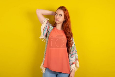 Téléchargez les photos : Femme aux cheveux roux debout sur fond jaune studio confondre et s'interroger sur la question. Incertain avec le doute, pensant la main sur la tête. Concept pensif. - en image libre de droit