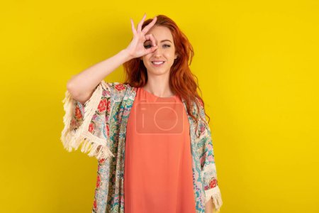 Téléchargez les photos : Femme aux cheveux rouges debout sur fond jaune studio faisant un geste correct avec le sourire de la main, les yeux regardant à travers les doigts avec un visage heureux. - en image libre de droit