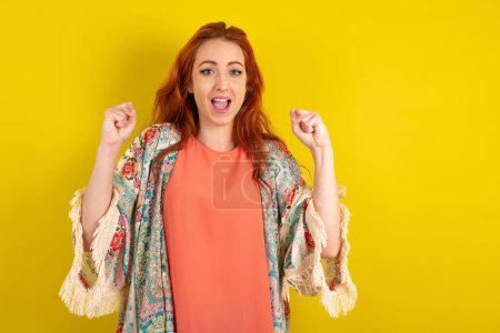 Photo for Red haired woman standing over yellow studio background celebrating surprised and amazed for success with arms raised and open eyes. Winner concept. - Royalty Free Image
